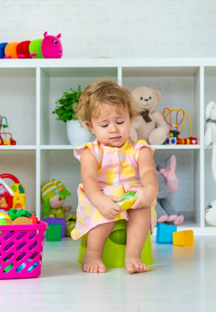 Toddler on a green potty holding a toy in a bright playroom
